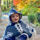 toddler sitting down with plants in the background and wearing a blue and white poncho with hood on