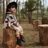 toddler sitting on a tree log wearing a black hat and a poncho with colors and a white pattern