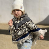toddler running wearing a gray and blue poncho and a white beanie 