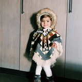 toddler with a closet behind her facing the camera with a poncho with colors and a hood with fur 