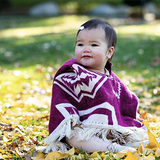 baby sitting down on the grass with dry leaves wearing a poncho color put and white  and looking to the side