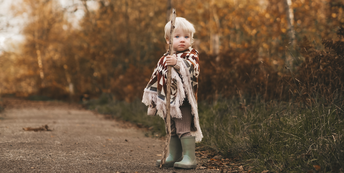 toddler standing in the distance in the woods, looking at the camera wearing a poncho and rainboots and holding a stick