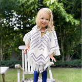 toddler sitting on top of a lounge chair in a garden wearing a lilac and white poncho and blue pants 