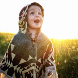 baby wearing a poncho with the hood on in a firld of flowers with the sun setting 