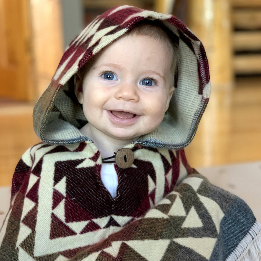 baby smiling looking at the camera with a poncho with the hood on with colors brown, pink, yellow and blue with a white patten on top