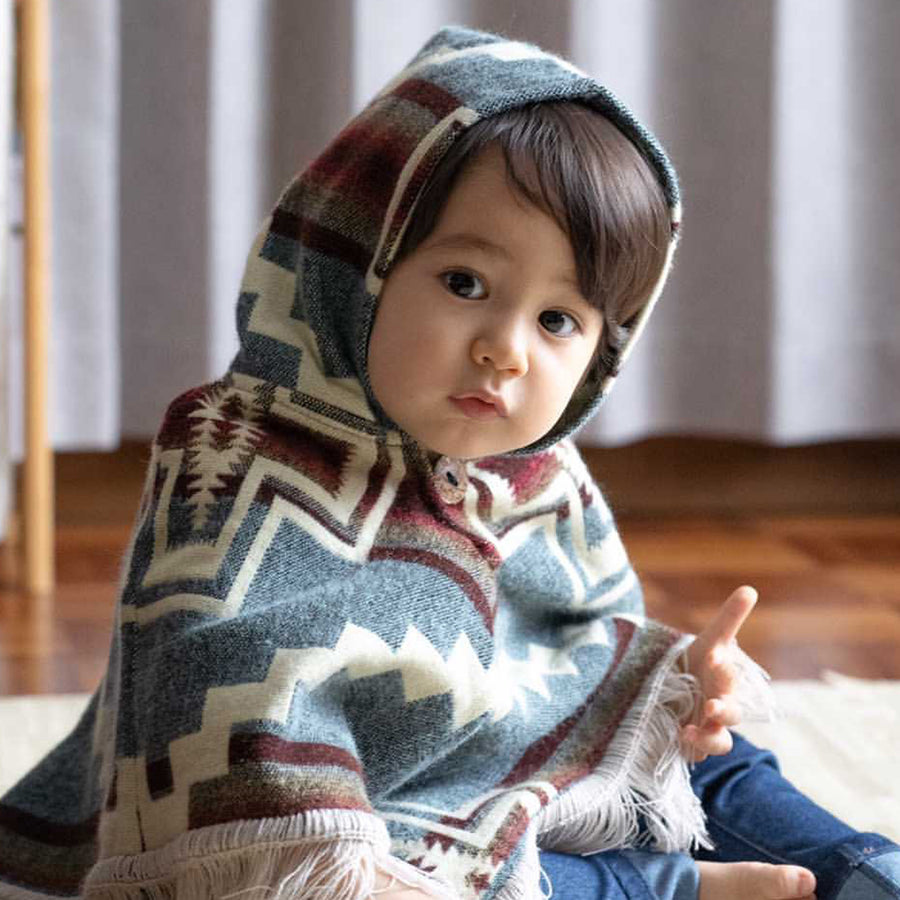 toddler sitting on the floor, looking at the camera wearing a light blue with orange stripes poncho with the hood on