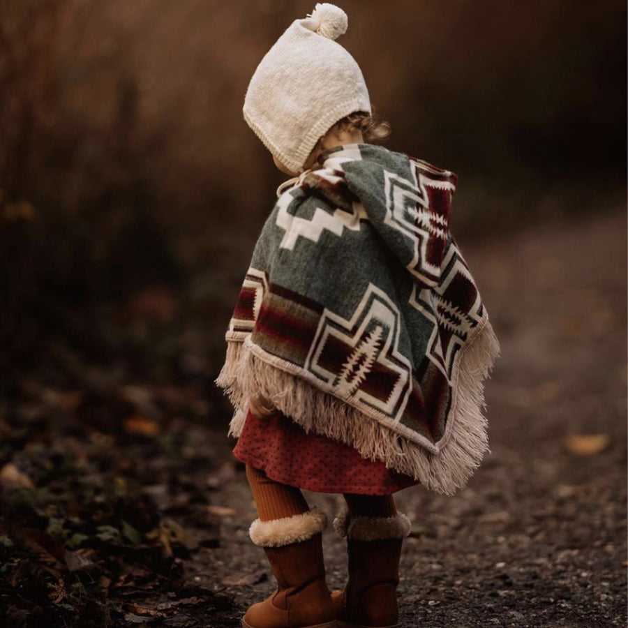 toddler standing facing away from the camera wearing a white beanie and a light blue with orange stripes poncho with fringe
