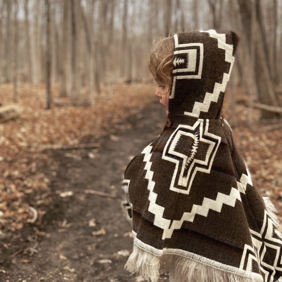toddler facing sideways to the left wearing a brown and white poncho with the hood oon