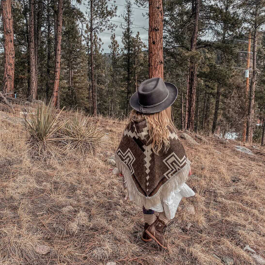 toddler walking away from the camera in the woods wearing a brown and white poncho and a black hat