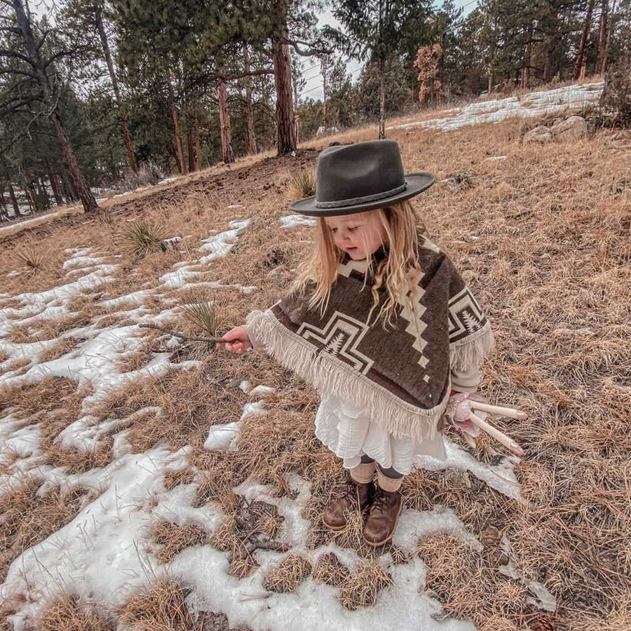 toddler looking at the floor playing with a stick wearing a brown and white poncho and a black hat