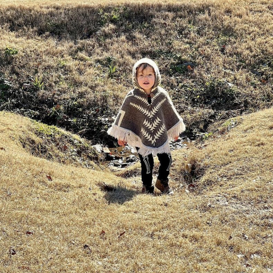 toddler looking at the camera in a field wear9ing a brown and white poncho with the hood on