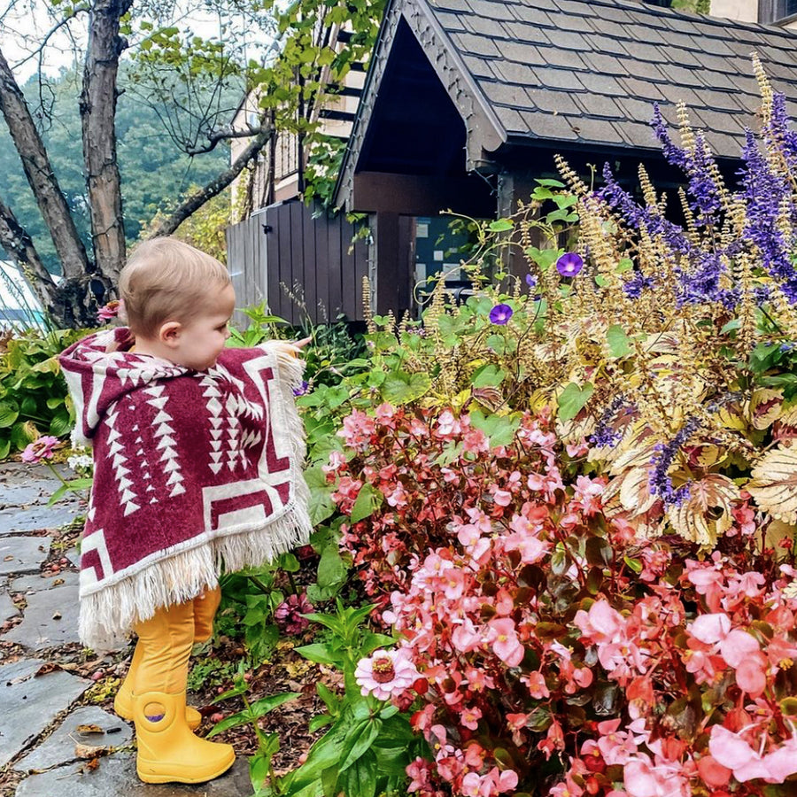 toddler outside playing with flowers wearing a plum and white poncho and yellow rain boots