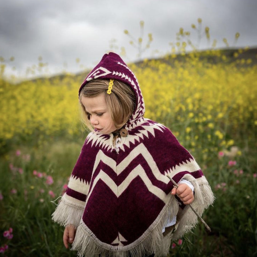 toddler in a flower field looking down wearing a plum and white poncho with the hood on and holding a stick