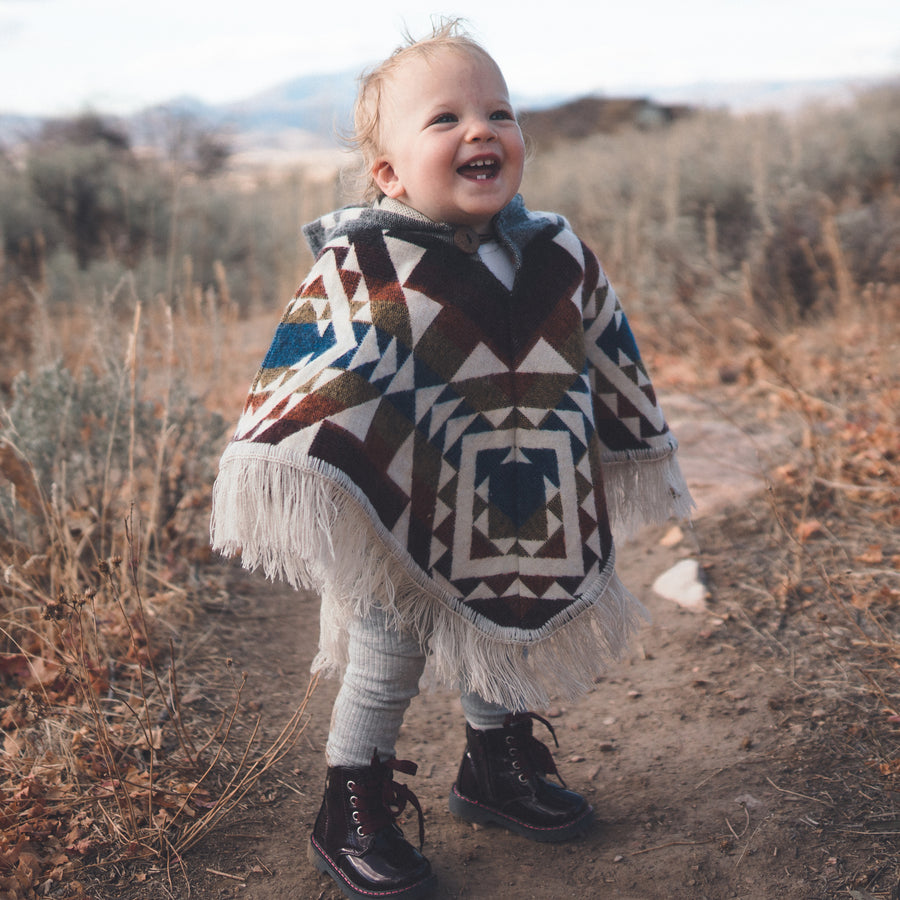 toddler smiling wearing a multicolor poncho with white patterns and black boots with a mountain background