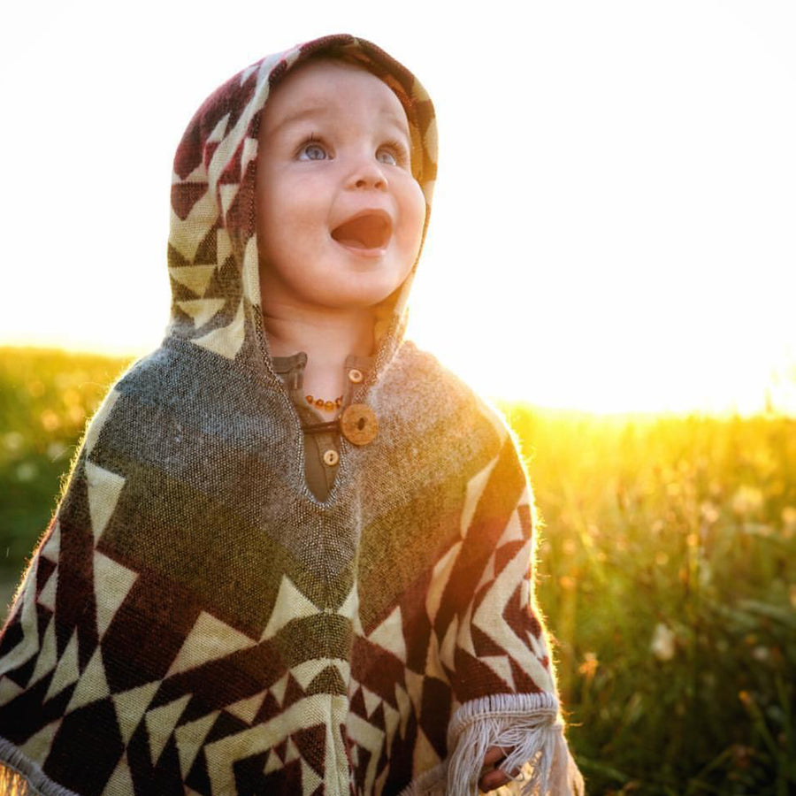 toddler in a field looking at the sky with mouth open wearing a poncho with reds, orange, yellow and light blue with white patterns and the hood on and the sun setting in the background