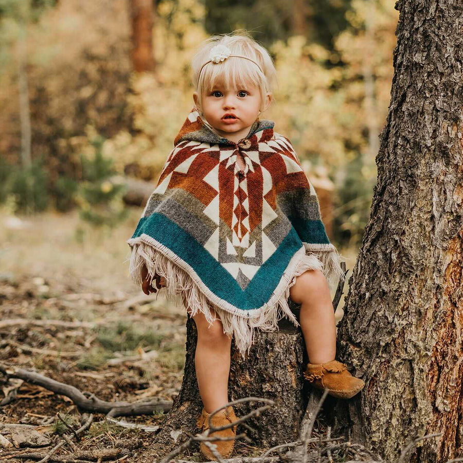 toddler sitting on a tree wearing a rainbow color poncho with white patterns and a flower headband on her hair and brown shoes