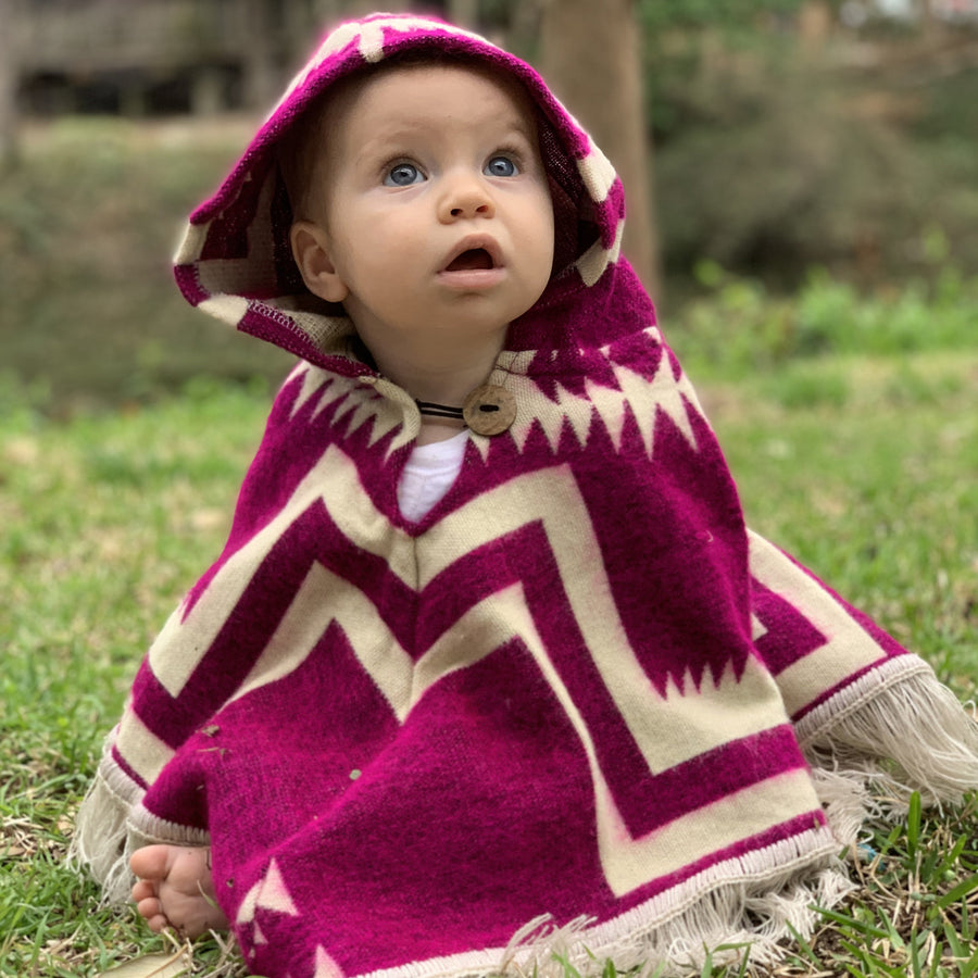 baby sitting on grass floor looking up wearing a plum and white color poncho with the hood on