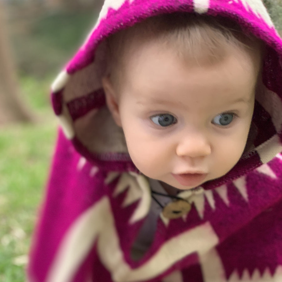 Closeup of baby wearing a plum color poncho with white details with the hood on