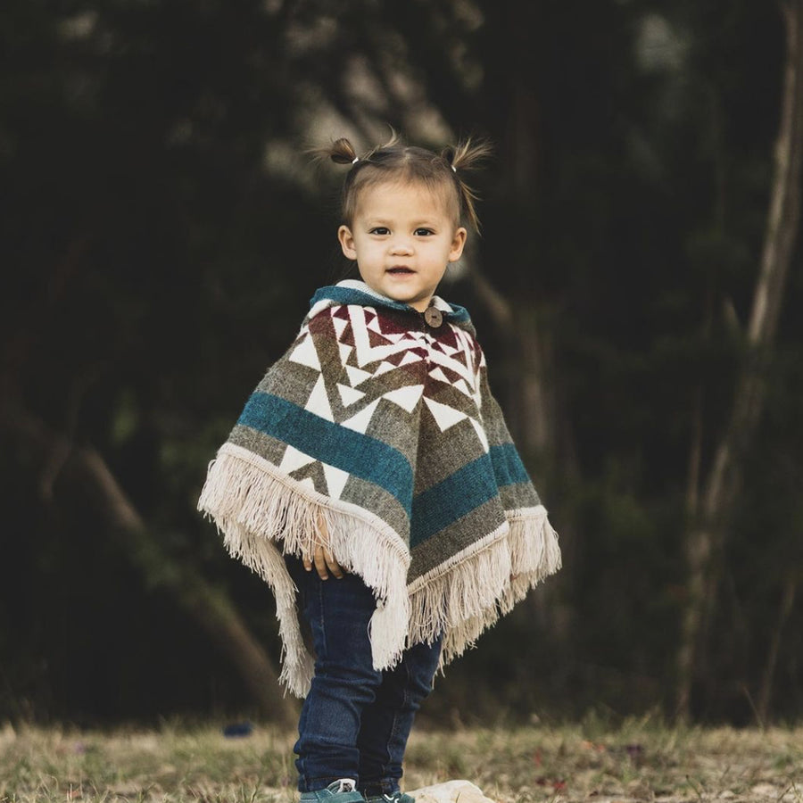toddler standing looking at the camera wearing a gray and turquouise poncho with red in the neck area ands white patterns 