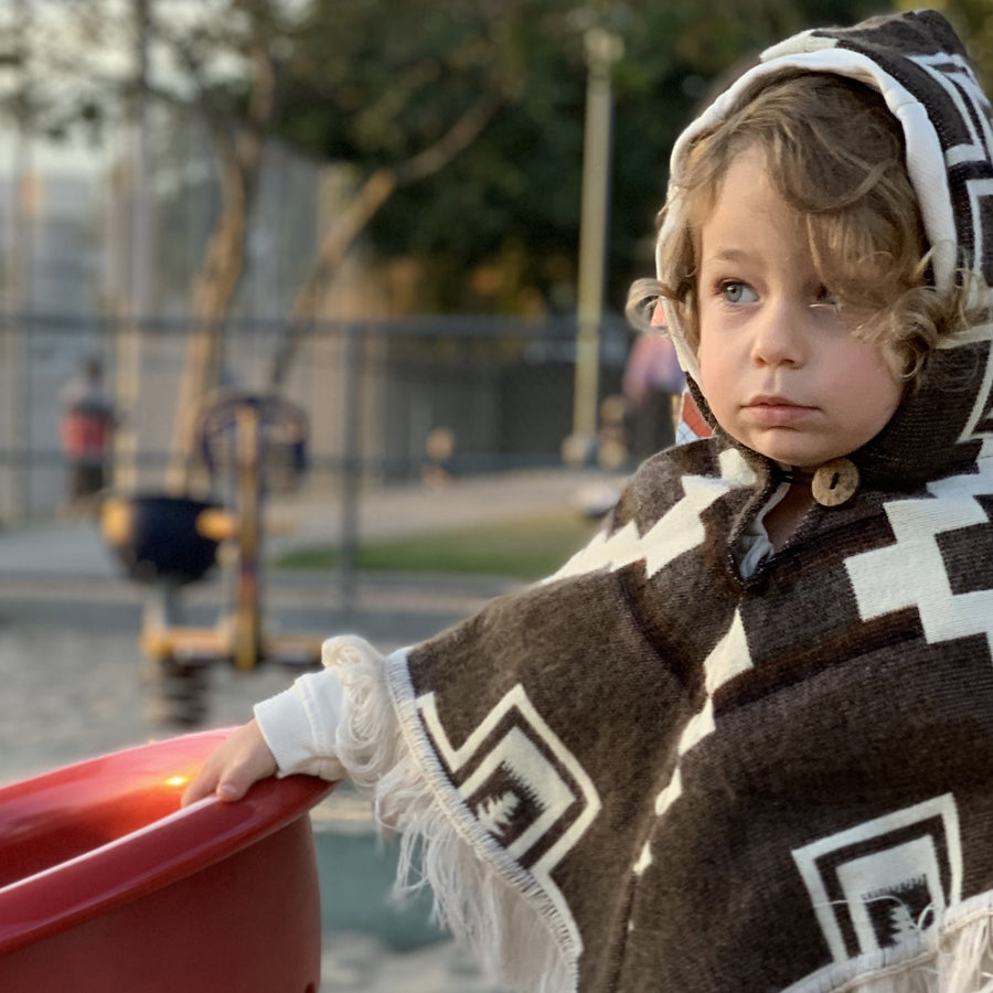 toddler at a playground wearing a brown poncho with white lines and crosses wearing the hood on 