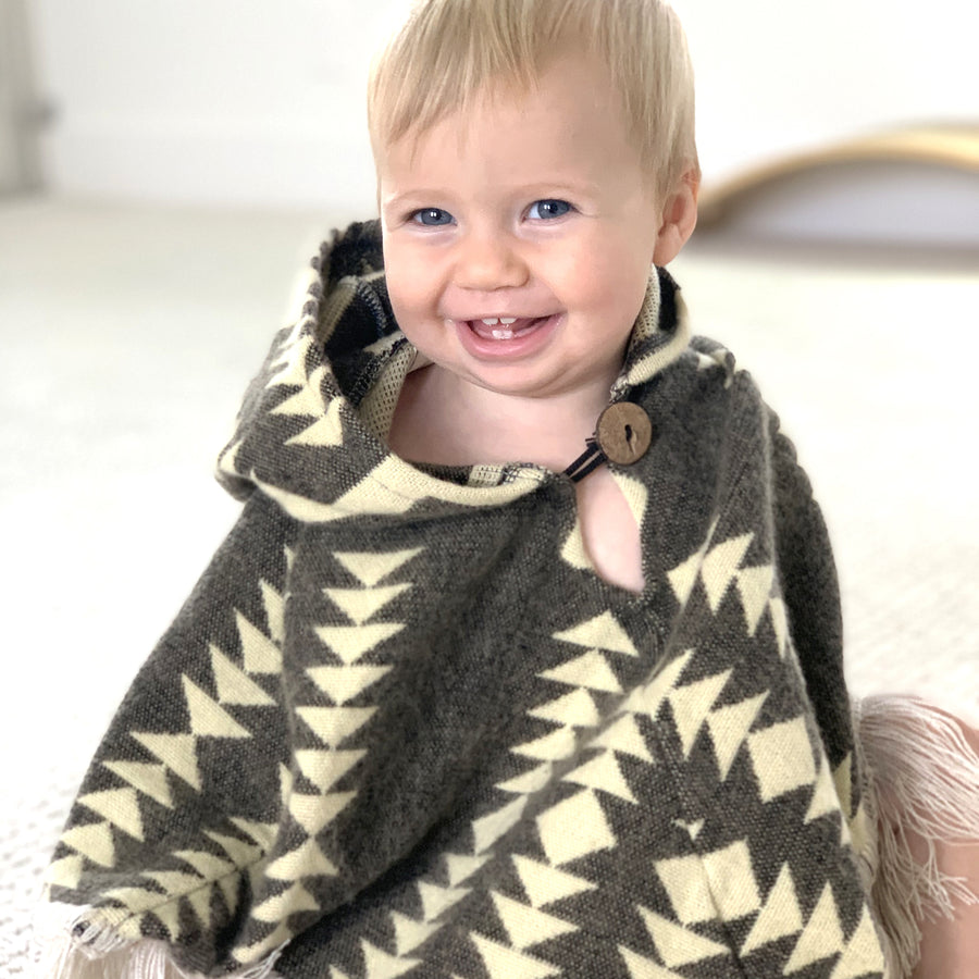baby smiling facing the camera sitting down wearing a baby poncho that is brown with cream color arrows. it has a button in the neck area and the hood in the back