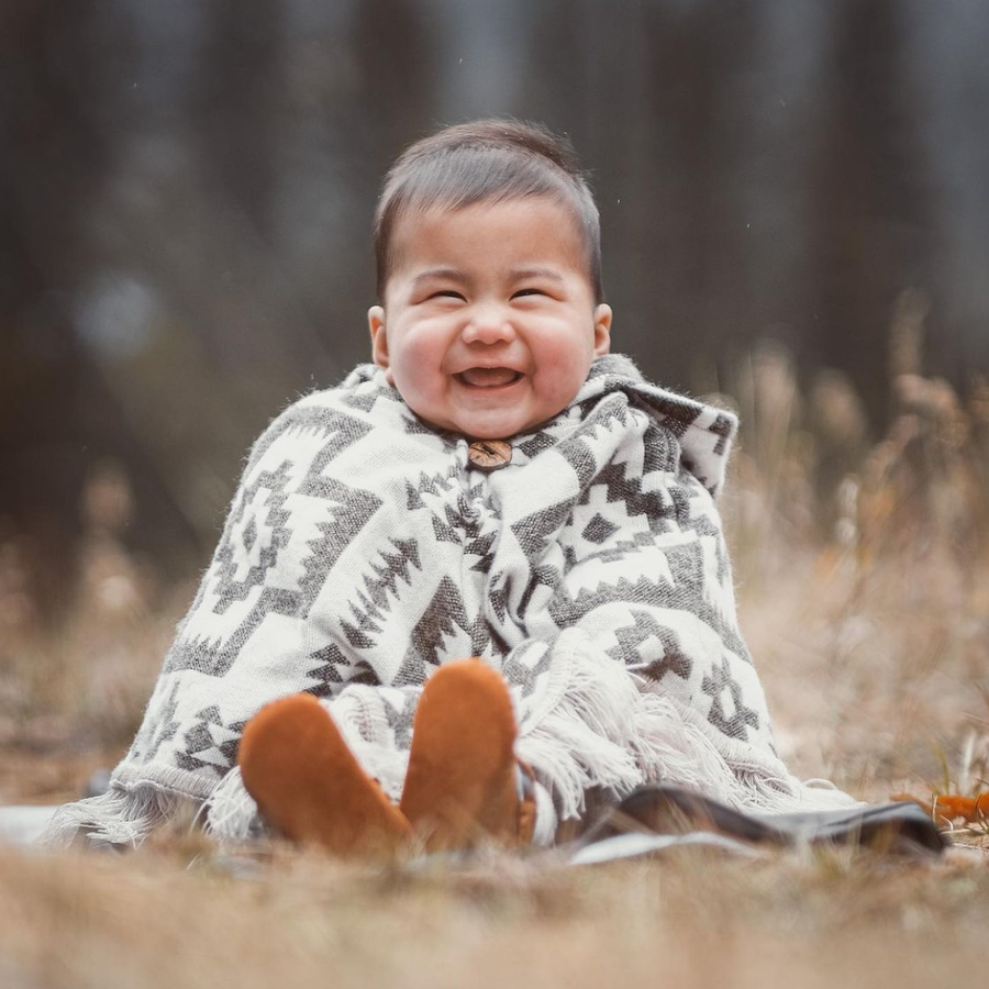 baby sitting down on dry grass, looking at the camera and smiling wearing a poncho with off white background and brown patterns