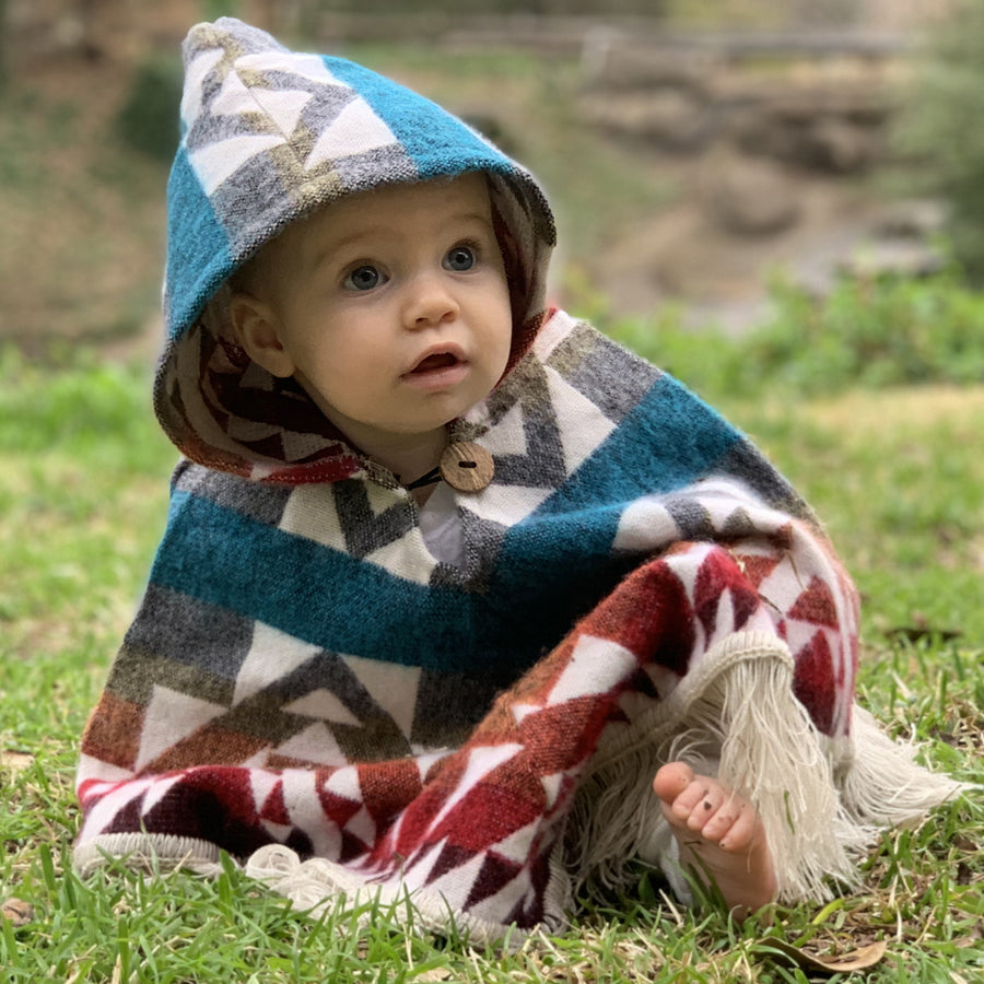 baby sitting on the floor wearing a  poncho with the hood on,  that is blue, gray, yellow, orange, red and dark red with white patterns on top