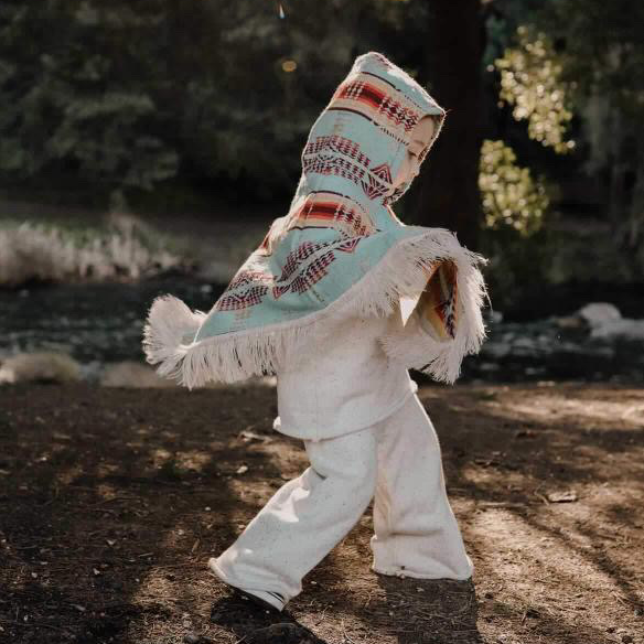 toddler running wearing white clothes and a light blue and orange and red stripes poncho with the hood on