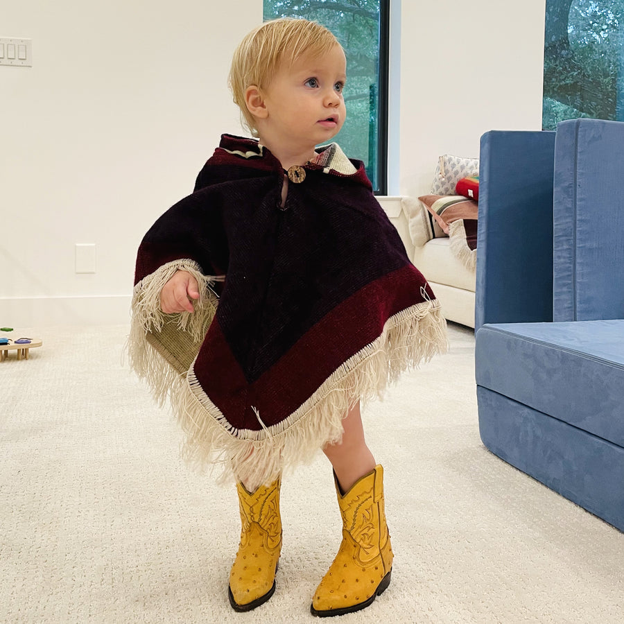toddler standing and looking to the right wearing a poncho with the hood down with colors purple and red/purple and fringe and wearing yellow cowboy boots