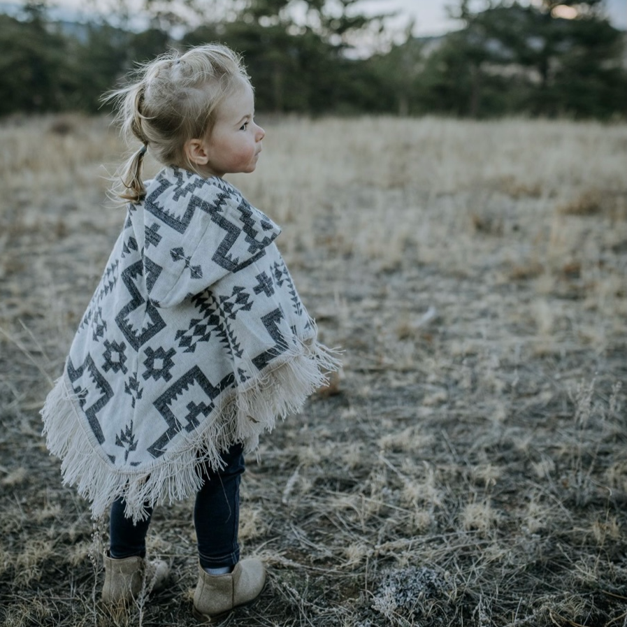 toddler standing facing away from the camera,  looking to the right wearing a white and lavender color poncho  