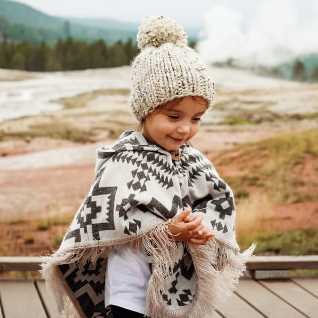 toddler standing looking down and grabbing her hands, wearing a poncho and a beanie