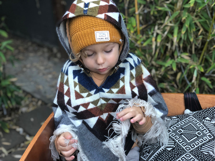 toddler sitting and looking down wearing a poncho with blue, yellow, orange, dark red and gray poncho with the hood on and a mustard color beanie